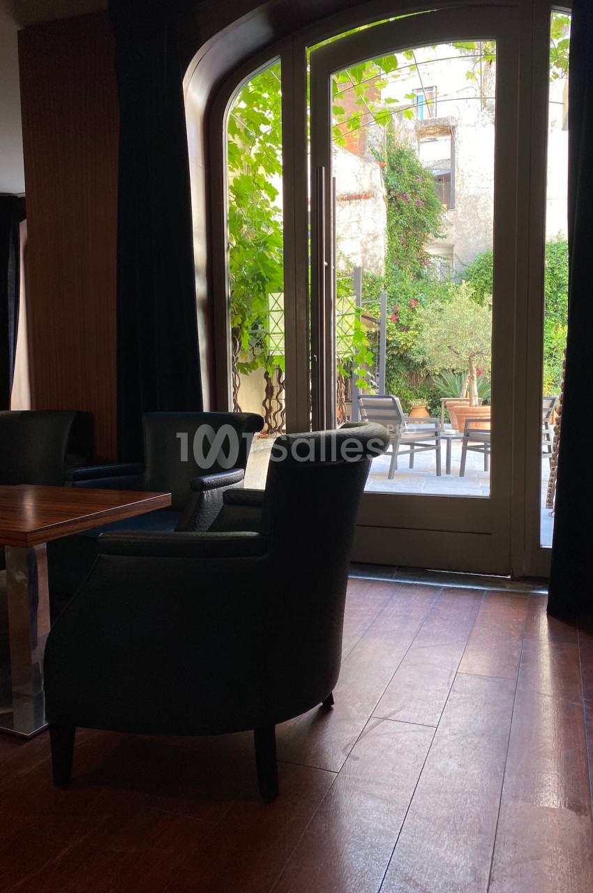 Espace intérieur avec fauteuils sombres près d'une porte vitrée donnant sur une terrasse verdoyante.