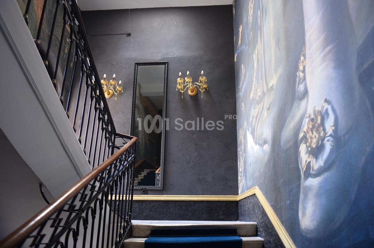 Escalier intérieur avec rampe en fer forgé, miroir mural, appliques dorées et fresque artistique sur le mur.