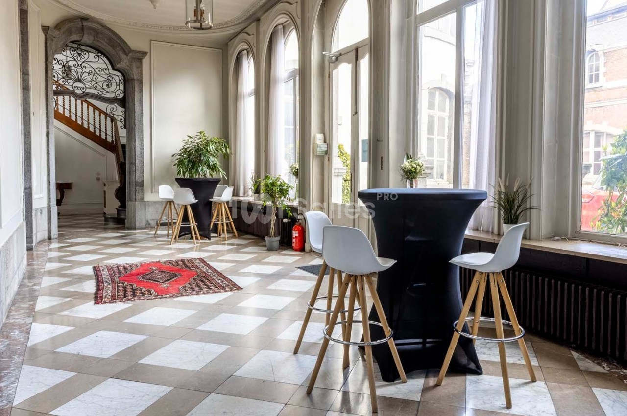 Salle lumineuse avec grandes fenêtres, sol en carreaux, tables hautes, chaises blanches et plantes décoratives.