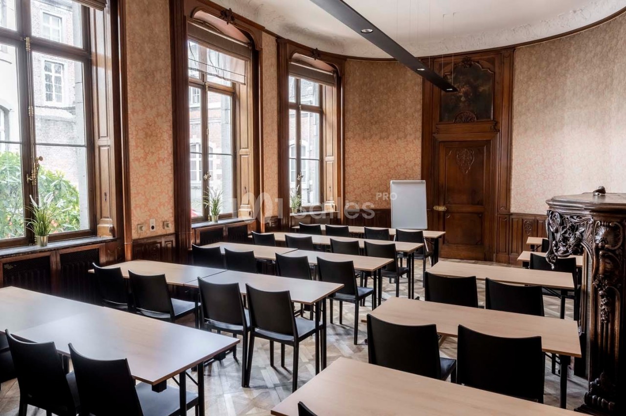 Salle de réunion lumineuse avec des tables en bois clair, des chaises noires et un tableau blanc devant des fenêtres.