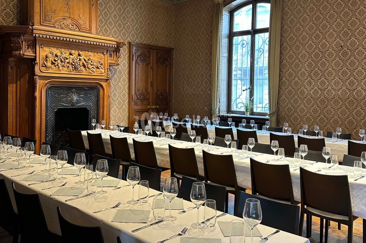 Salle élégante avec de longues tables dressées, nappes blanches, verres et couverts, devant une cheminée sculptée.