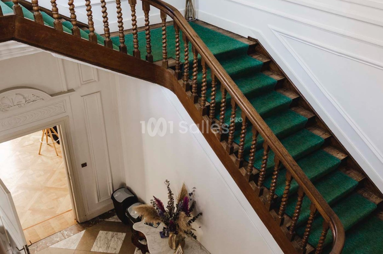 Escalier en bois avec tapis vert, balustrade sculptée, dans un intérieur lumineux avec murs blancs et décorations florales.