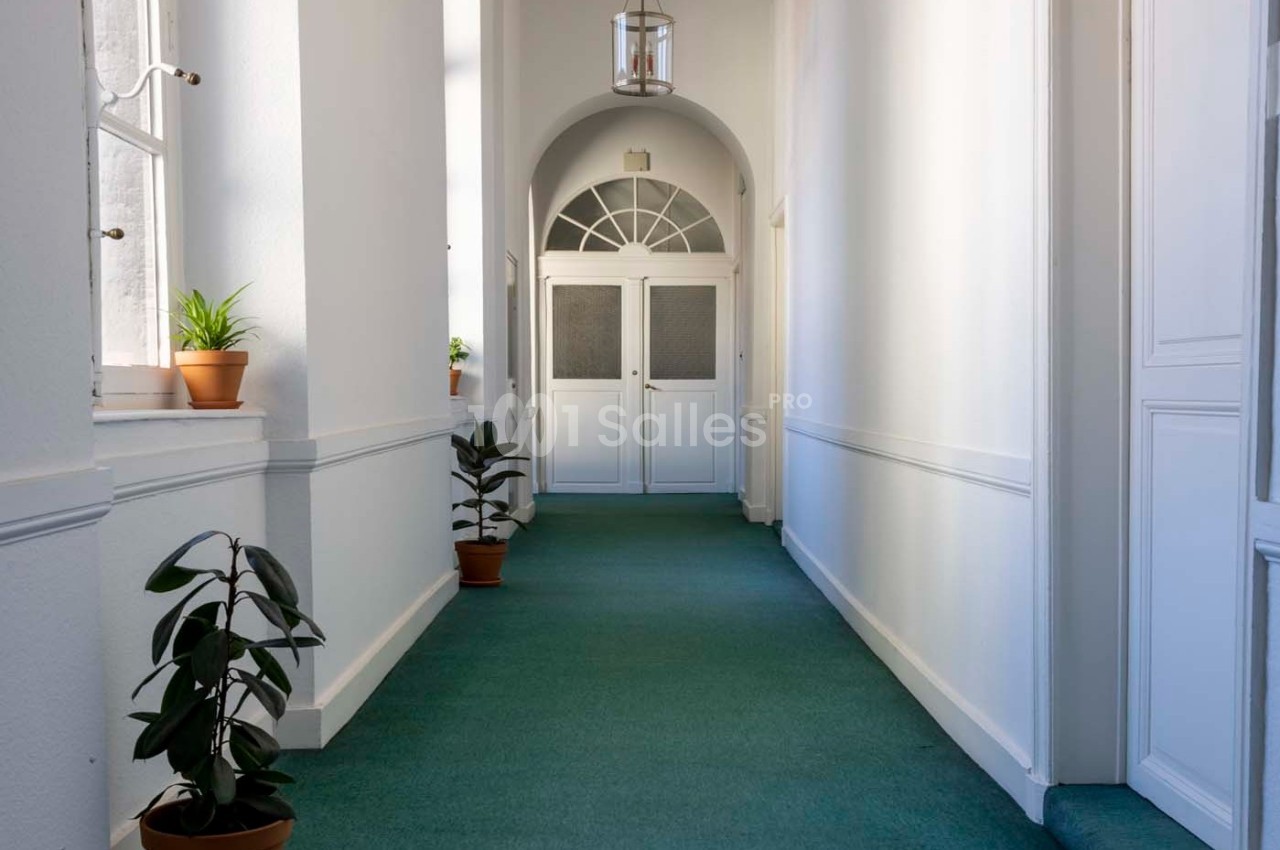 Couloir lumineux avec moquette verte, plantes en pot et portes blanches vitrées au fond.