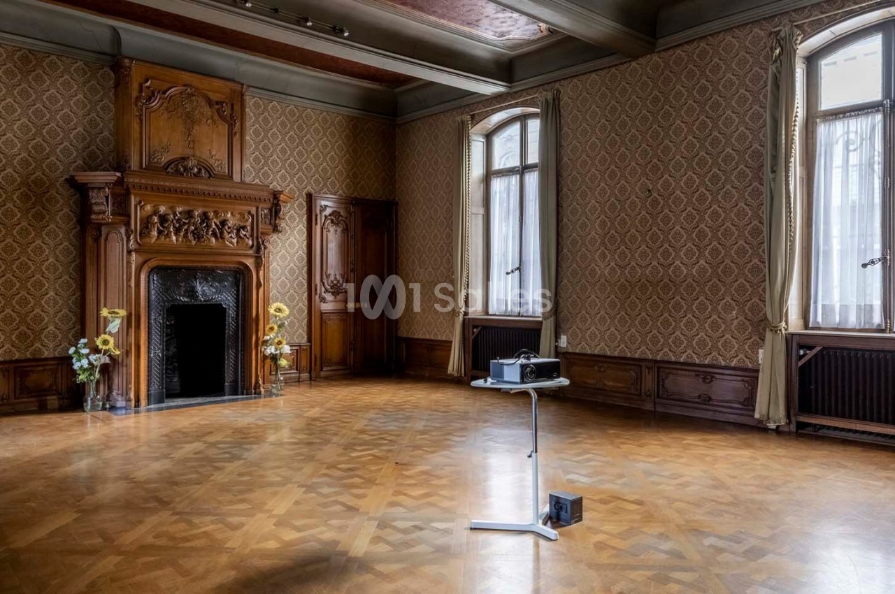 Salle ancienne avec parquet en bois, cheminée sculptée, murs ornés de motifs et un projecteur sur pied au centre.