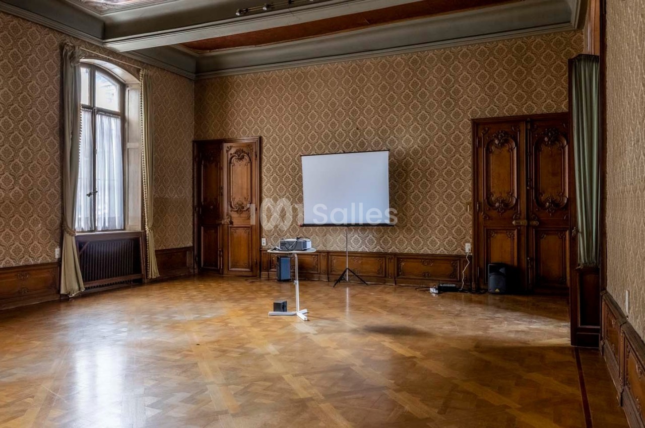 Salle vide avec parquet, murs ornés de motifs, grandes fenêtres, rideaux et un écran de projection installé.