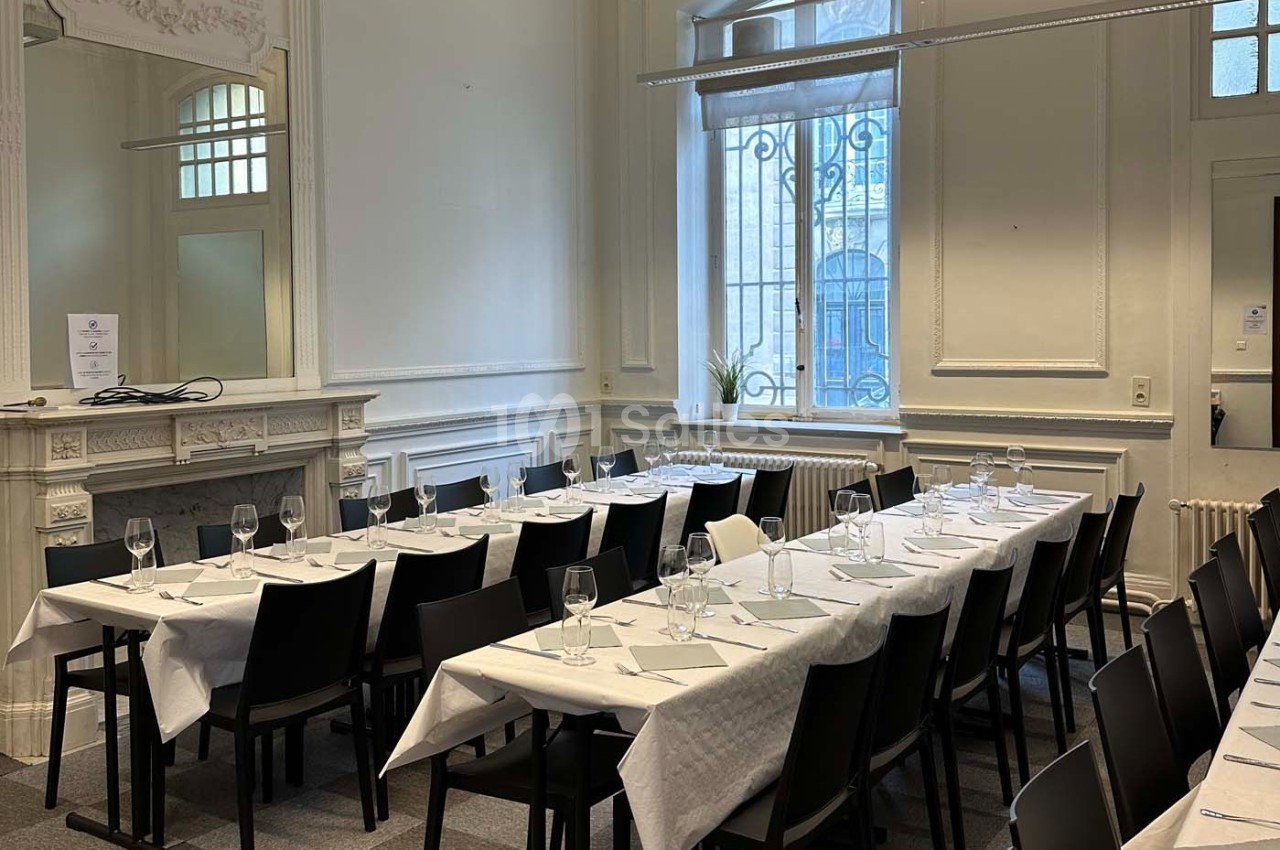 Salle lumineuse avec tables alignées, nappes blanches, verres et couverts, décorée de moulures et d'une cheminée.