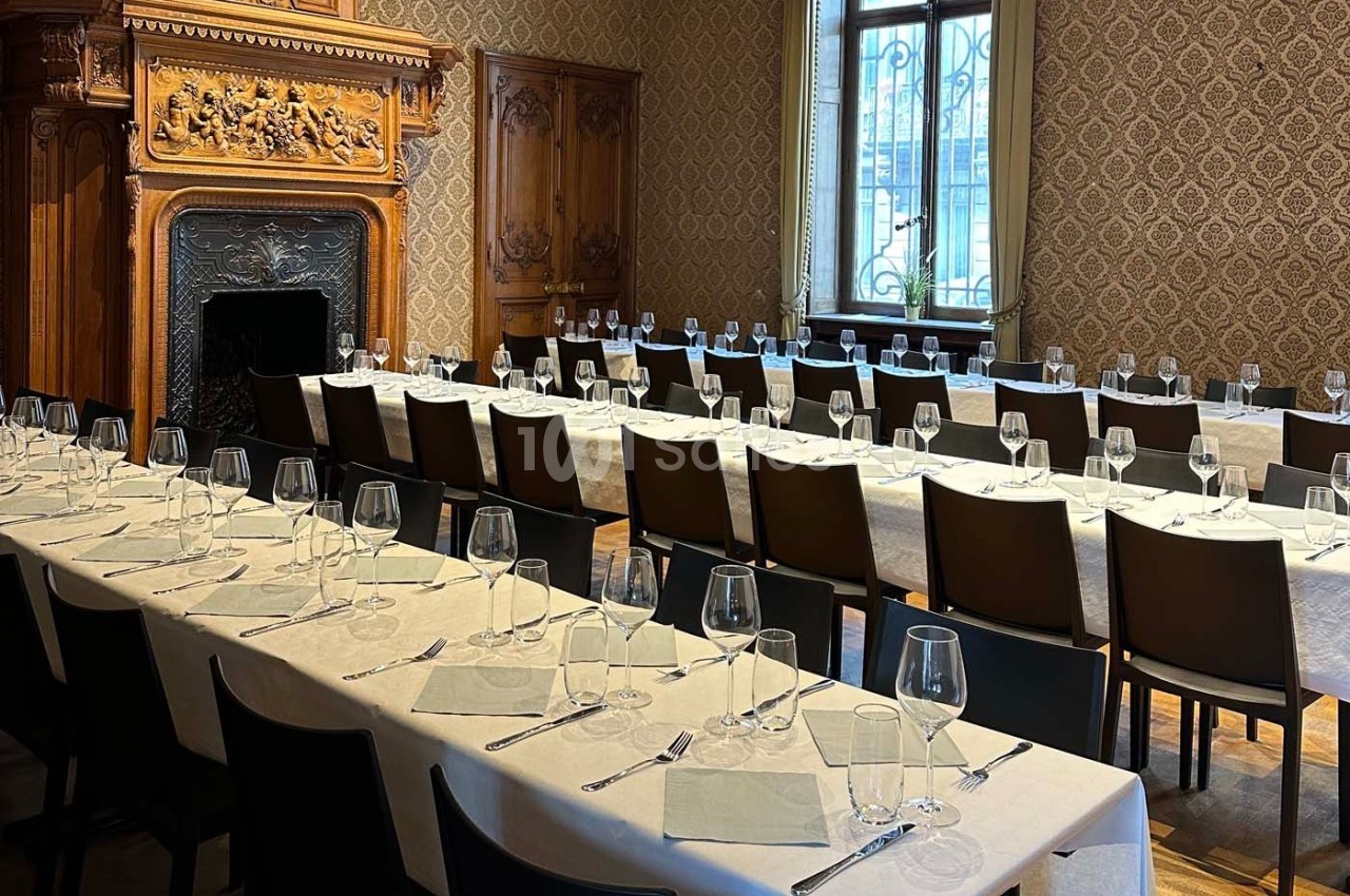 Salle élégante avec tables alignées, nappes blanches, couverts et verres, décorée de boiseries et d'une cheminée sculptée.