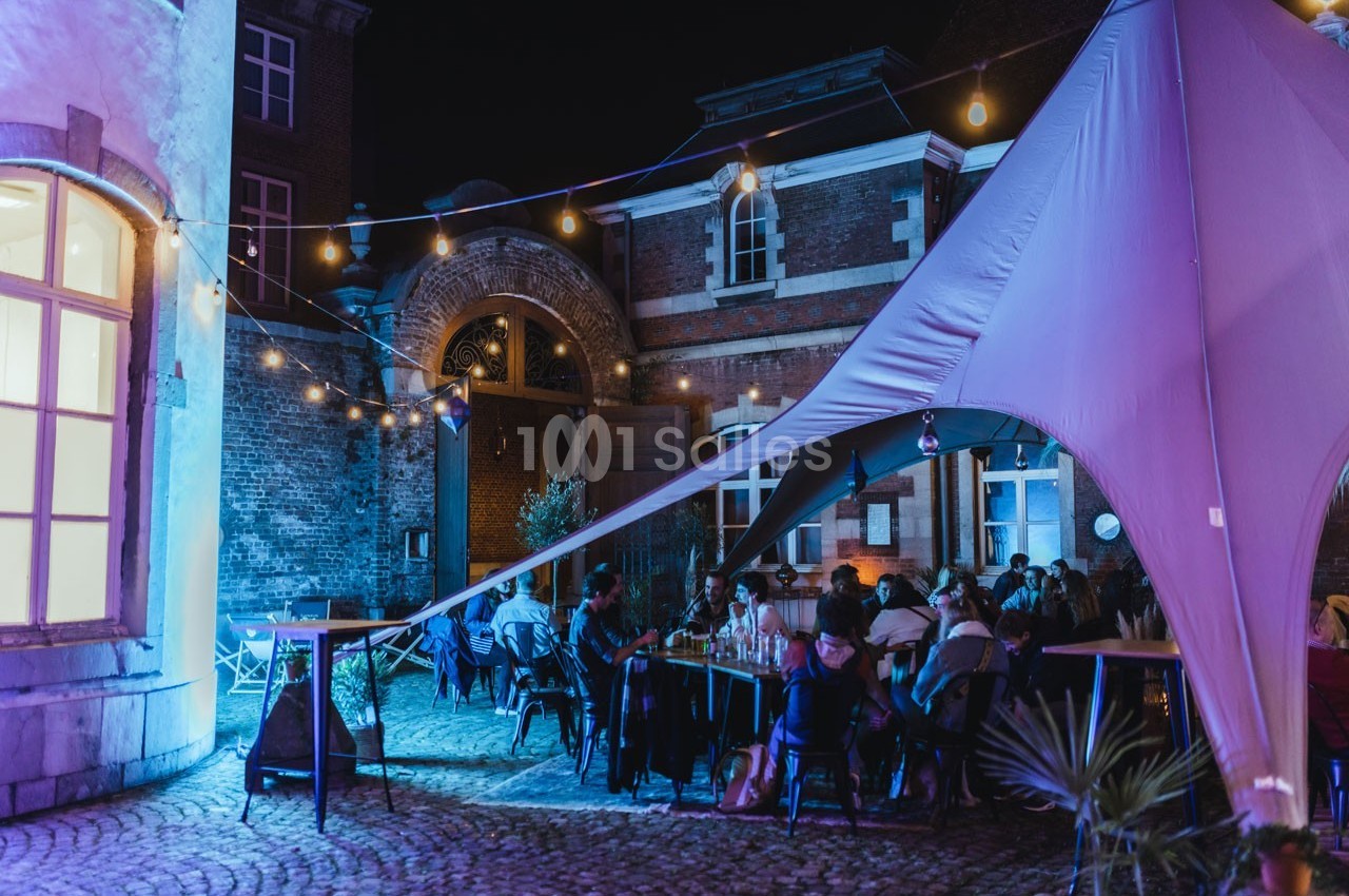 Cour intérieure éclairée avec des guirlandes lumineuses, des personnes assises sous une tente lors d'un événement nocturne.