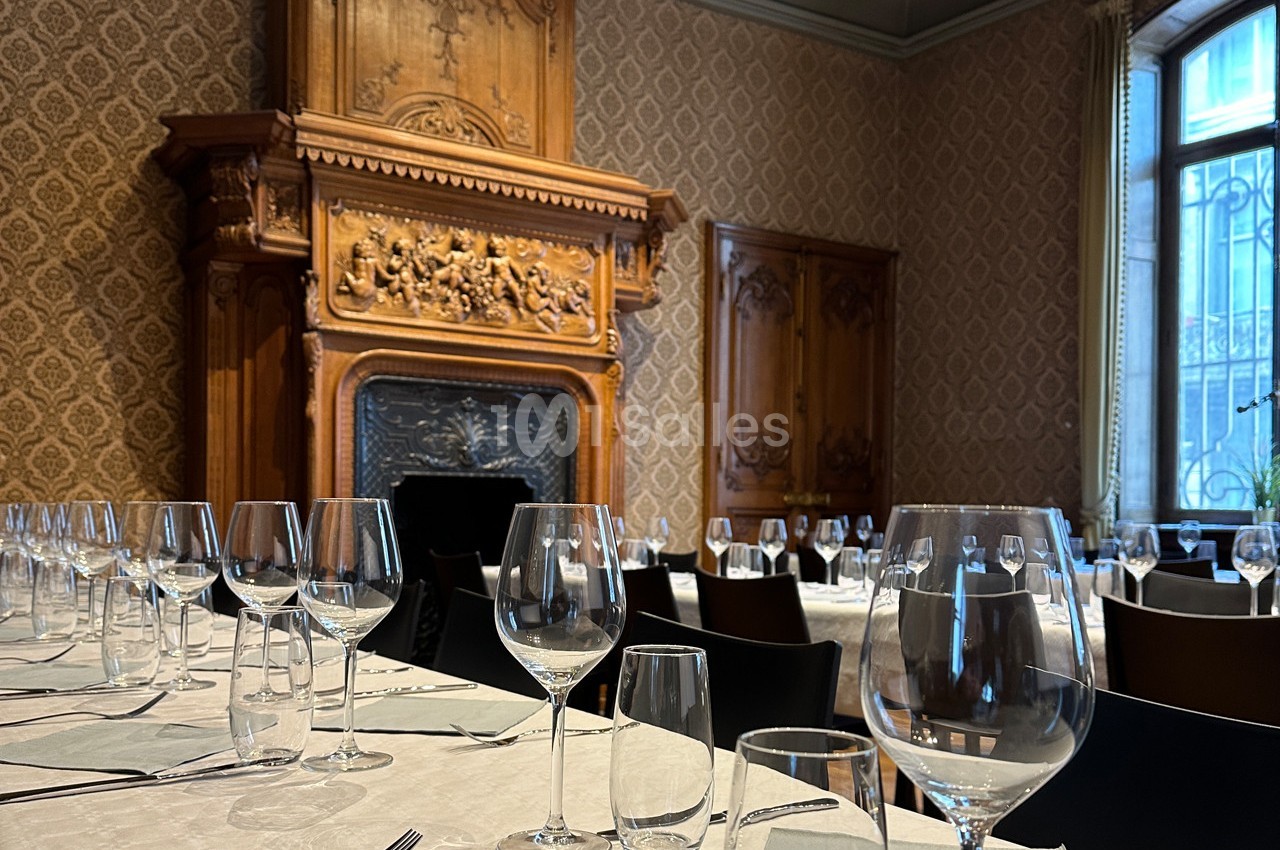 Salle à manger élégante avec cheminée sculptée en bois, tables dressées avec nappes blanches et verres alignés.