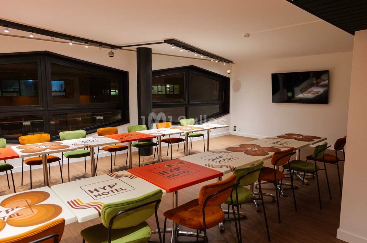 Salle de réunion lumineuse avec des tables colorées alignées, des chaises variées et un écran mural.