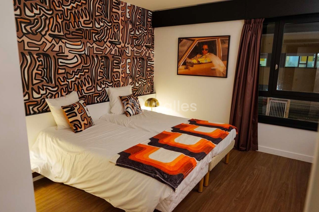 Chambre avec deux lits simples, décorée de motifs géométriques colorés et éclairée par une lampe de chevet dorée.