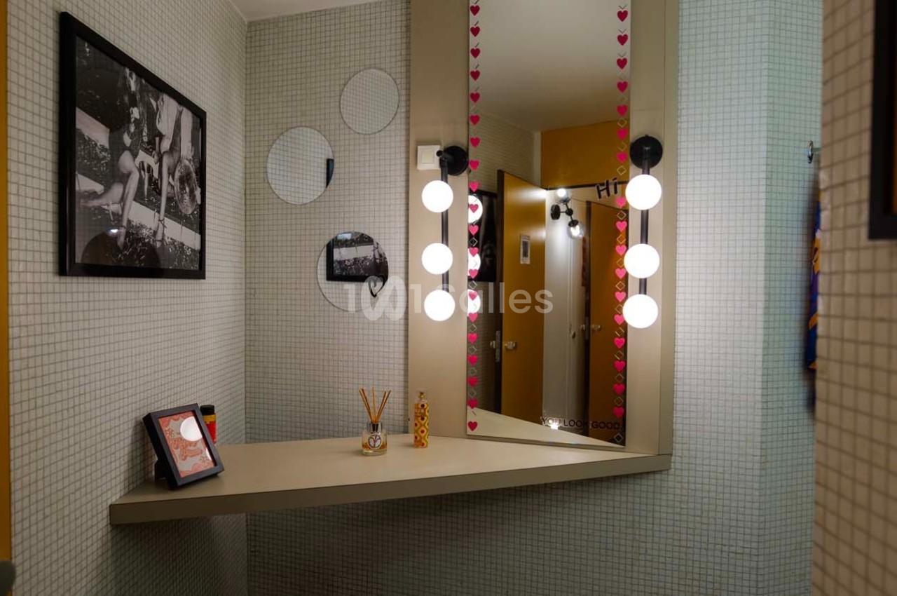 Miroir éclairé avec des ampoules, étagère décorée d'objets et murs recouverts de carreaux blancs.