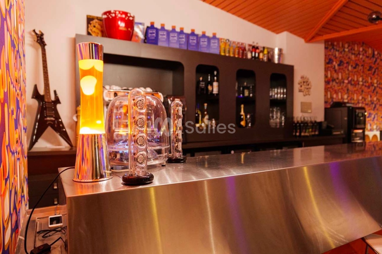 Bar avec comptoir en métal, lampe à lave, décorations colorées et étagères contenant des bouteilles à l’arrière.