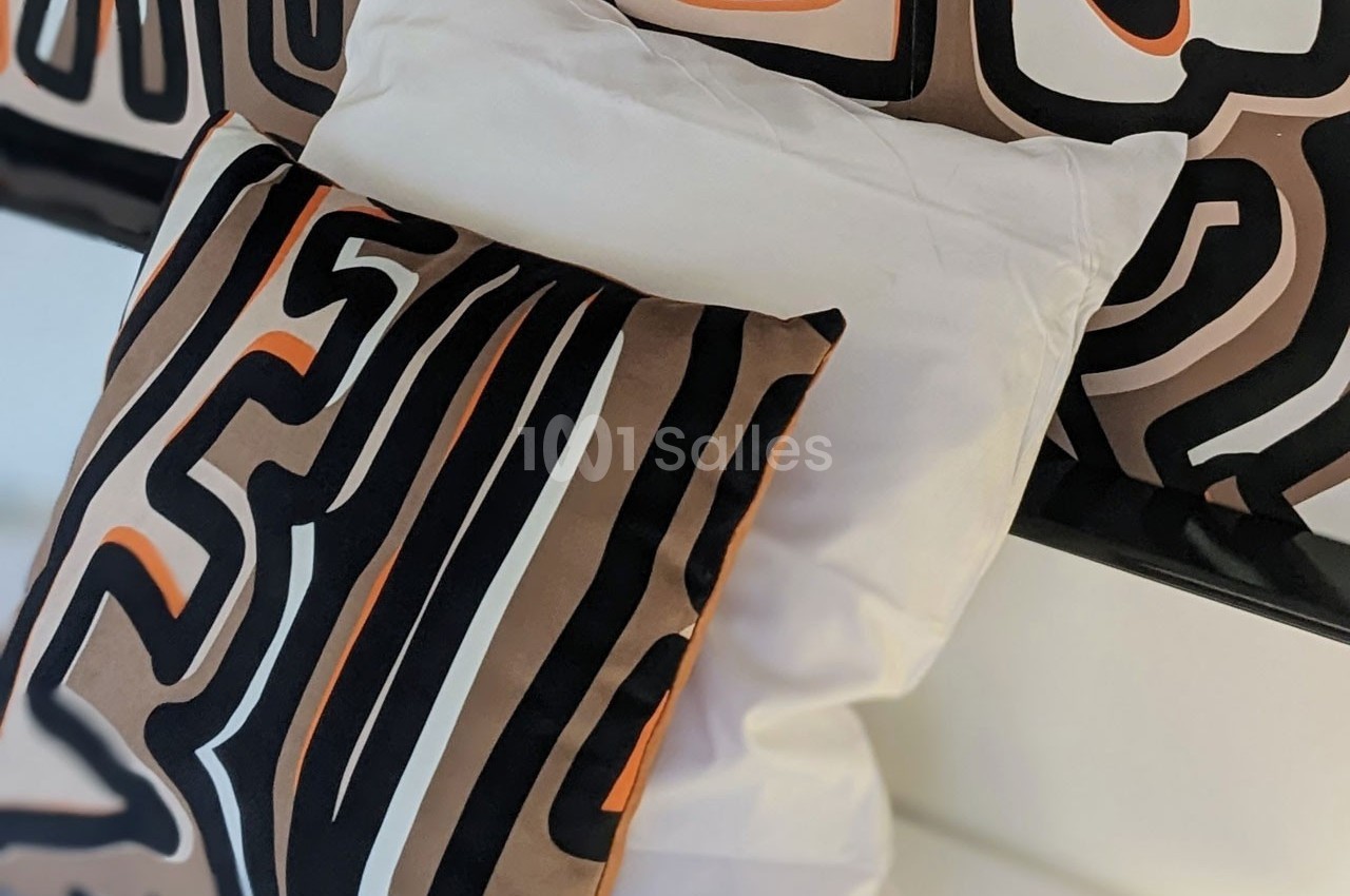 Coussins décoratifs avec motifs abstraits noirs, bruns et orange, posés sur un lit avec une housse blanche.