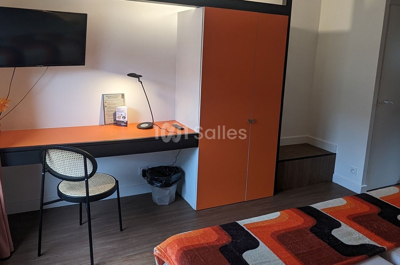 Bureau avec chaise, lampe, télévision murale et placard orange dans une chambre avec lit et couvre-lit coloré.