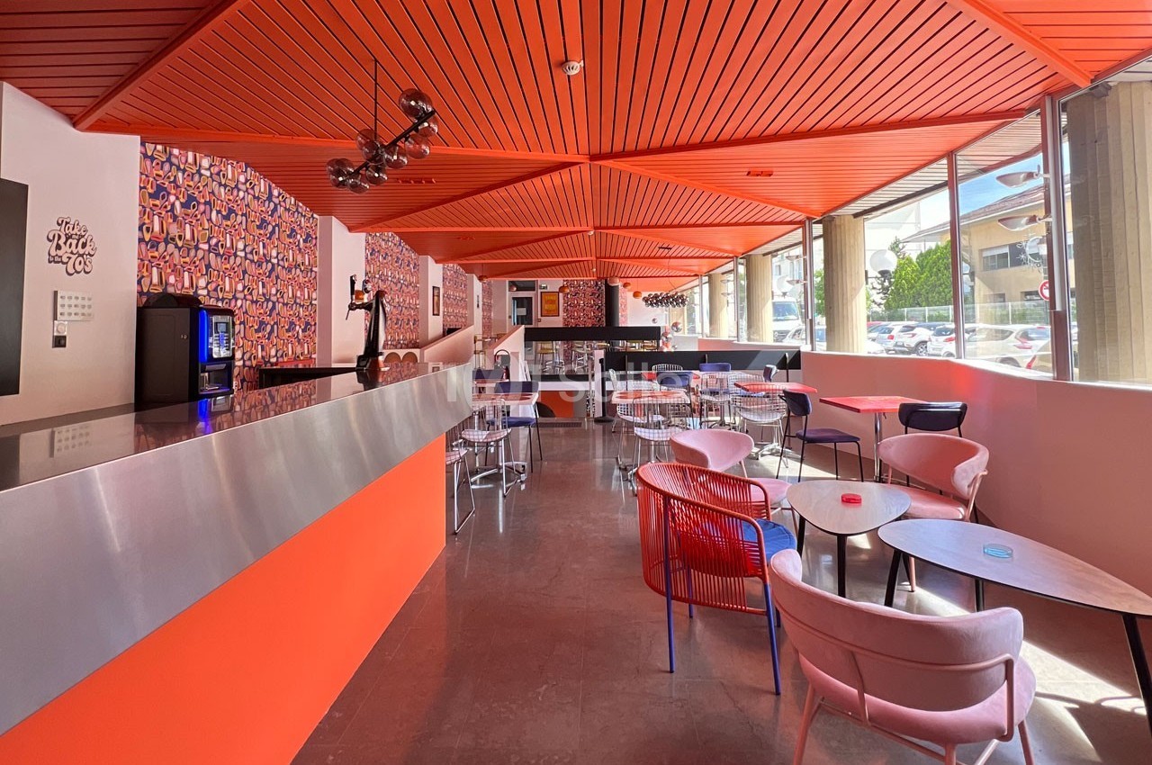 Salle lumineuse avec un bar moderne, des tables et des chaises colorées, et un plafond orange géométrique.