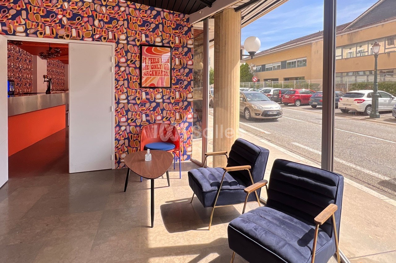 Espace d'attente lumineux avec fauteuils bleus, table basse en bois et mur coloré, vue sur la rue à travers une baie vitrée.