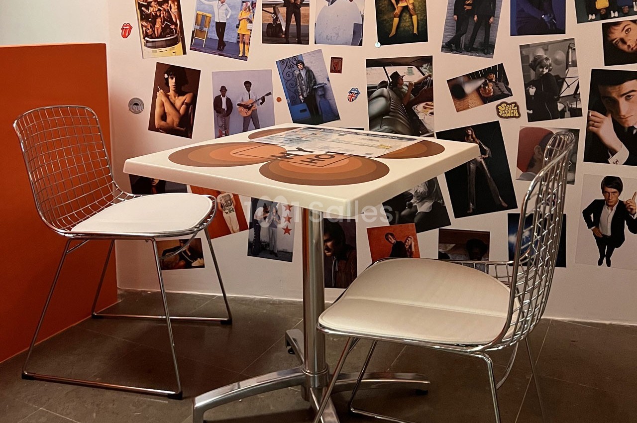 Deux chaises métalliques entourent une table carrée dans un coin décoré de photos murales variées.