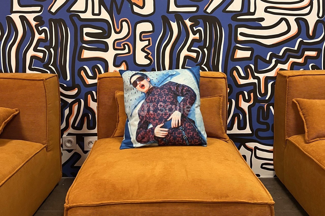 Fauteuil en velours orange avec un coussin imprimé d'un portrait, devant un mur décoré de motifs abstraits colorés.