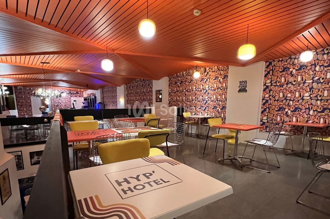 Salle de restaurant moderne avec tables colorées, chaises variées et plafond en bois éclairé par des suspensions.