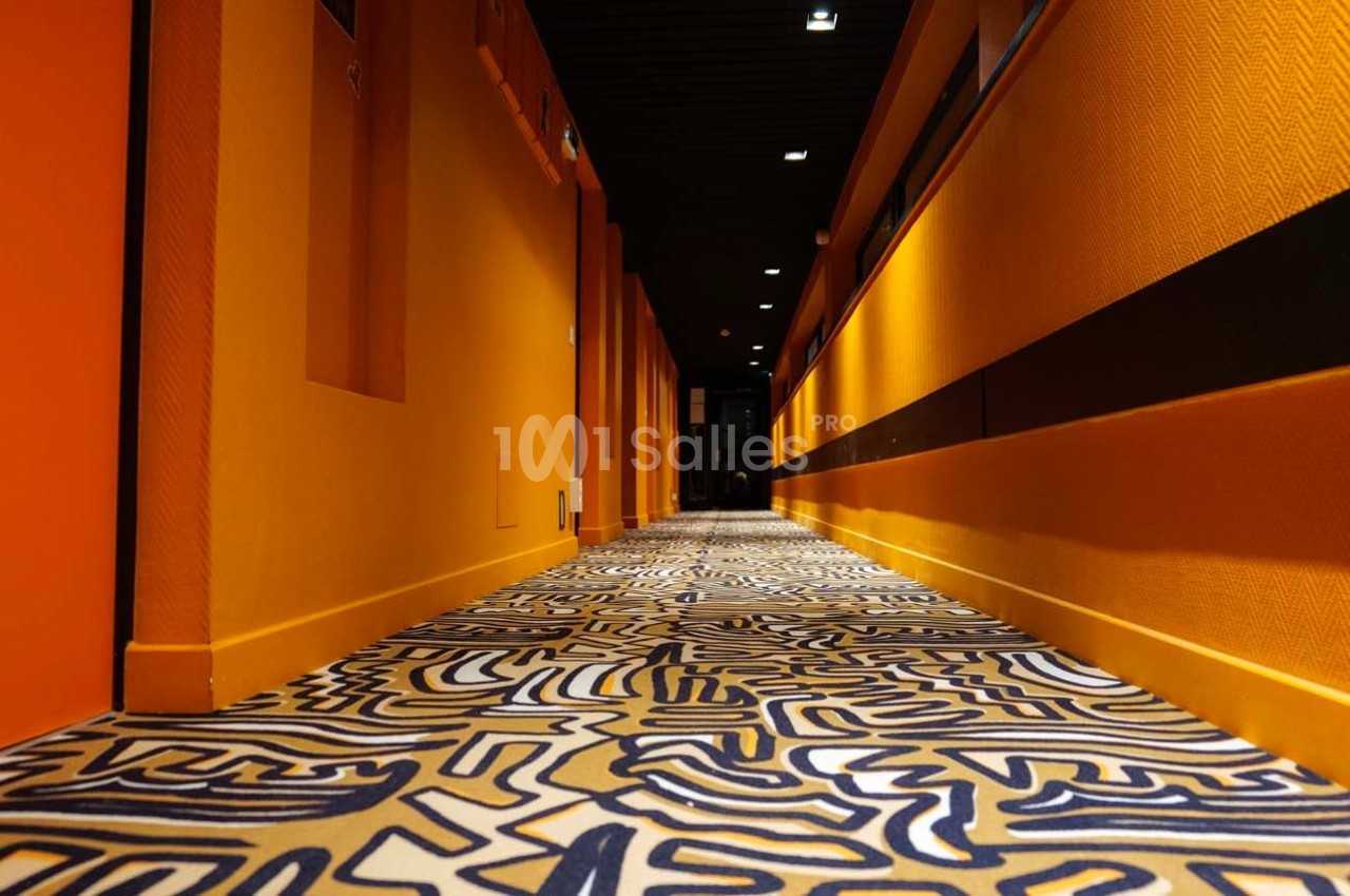 Couloir aux murs orange vif avec un sol à motifs abstraits colorés, éclairé par des lumières encastrées au plafond.