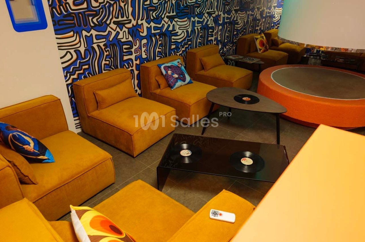 Salon avec fauteuils jaunes, table basse noire avec des vinyles, mur décoré de motifs abstraits bleus et blancs.