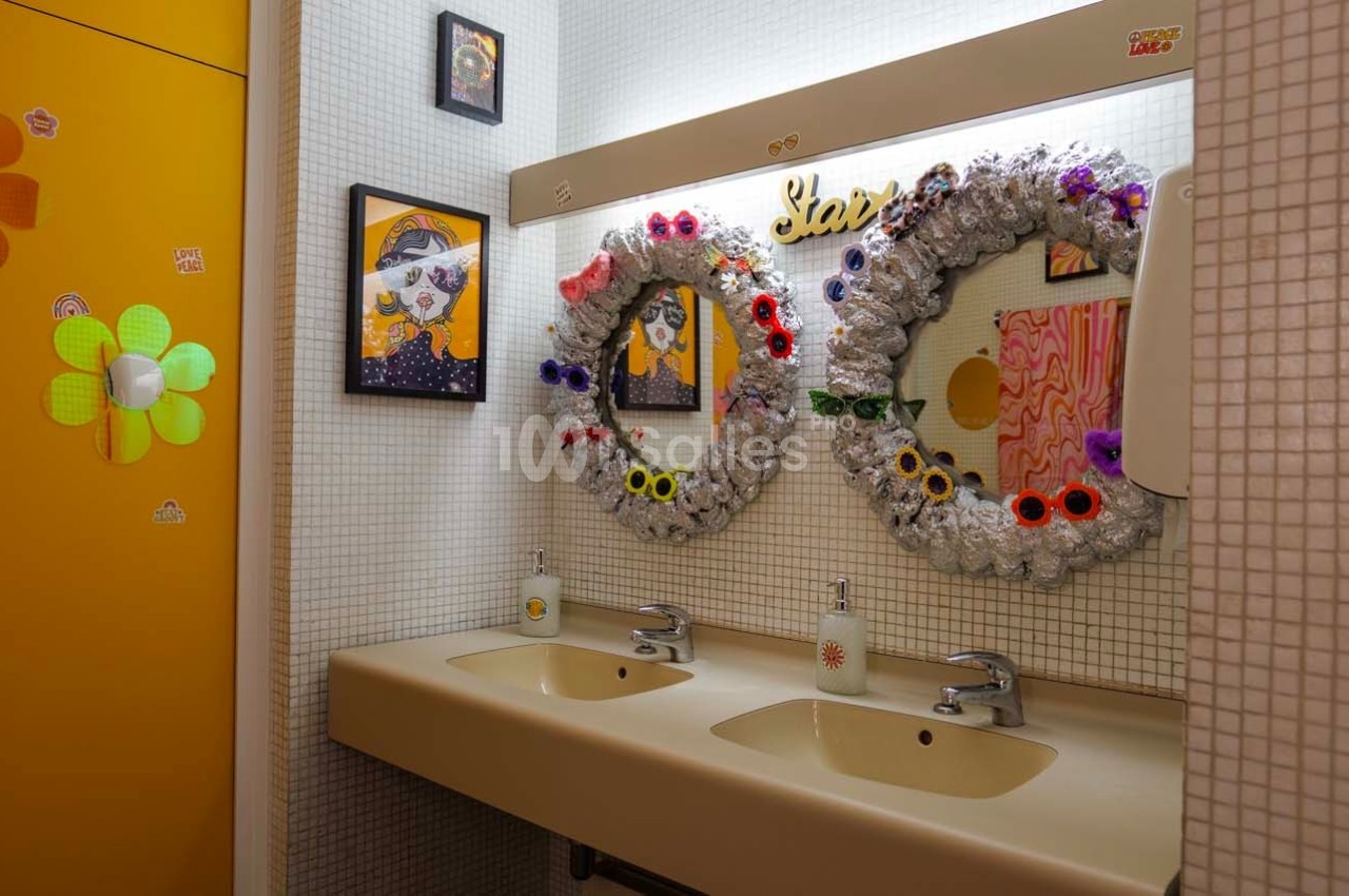 Deux lavabos avec miroirs décorés de couronnes colorées, entourés de cadres et d'un mur carrelé blanc.