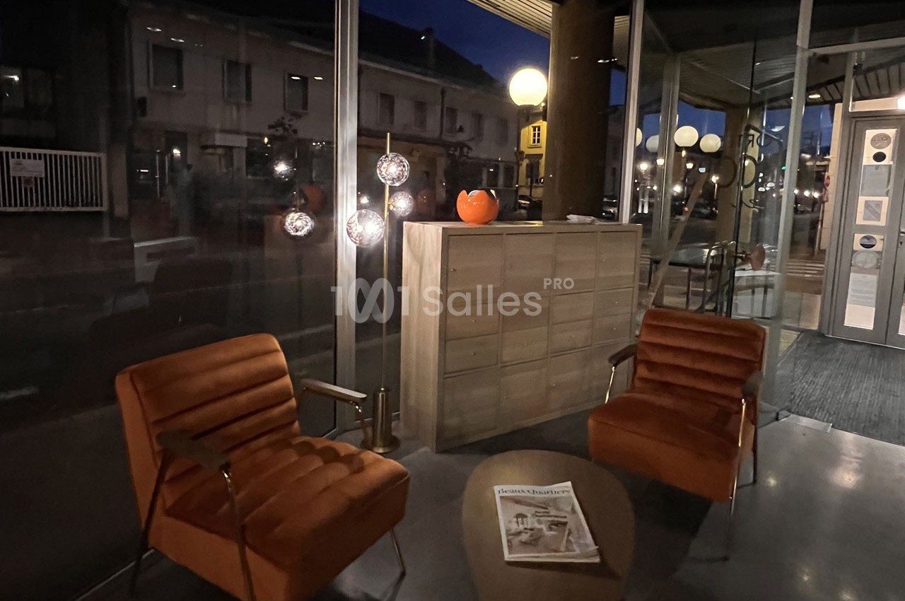 Deux fauteuils orange et une table basse avec un journal dans un espace vitré éclairé par des lampes en soirée.
