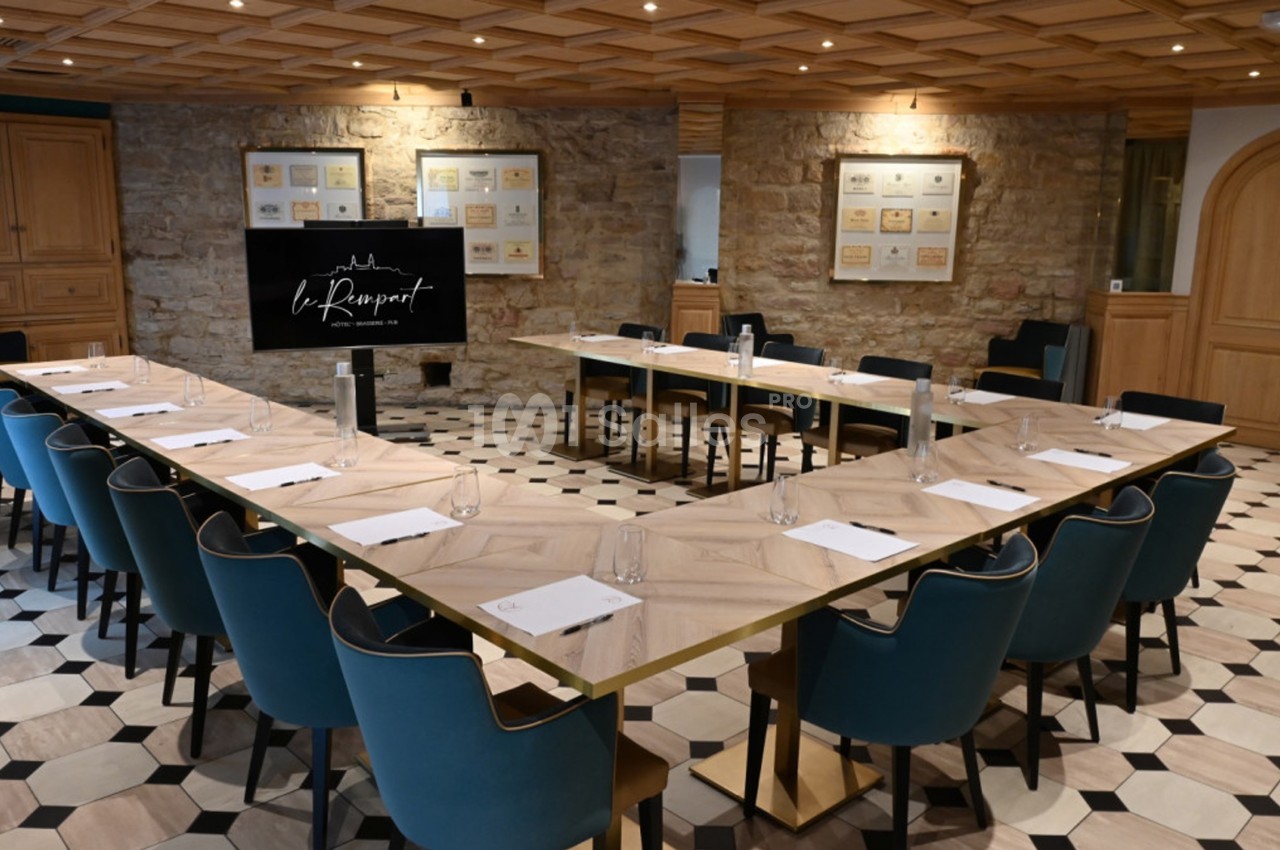 Salle de réunion avec tables en U, chaises bleues, écran de présentation et murs en pierre apparente.