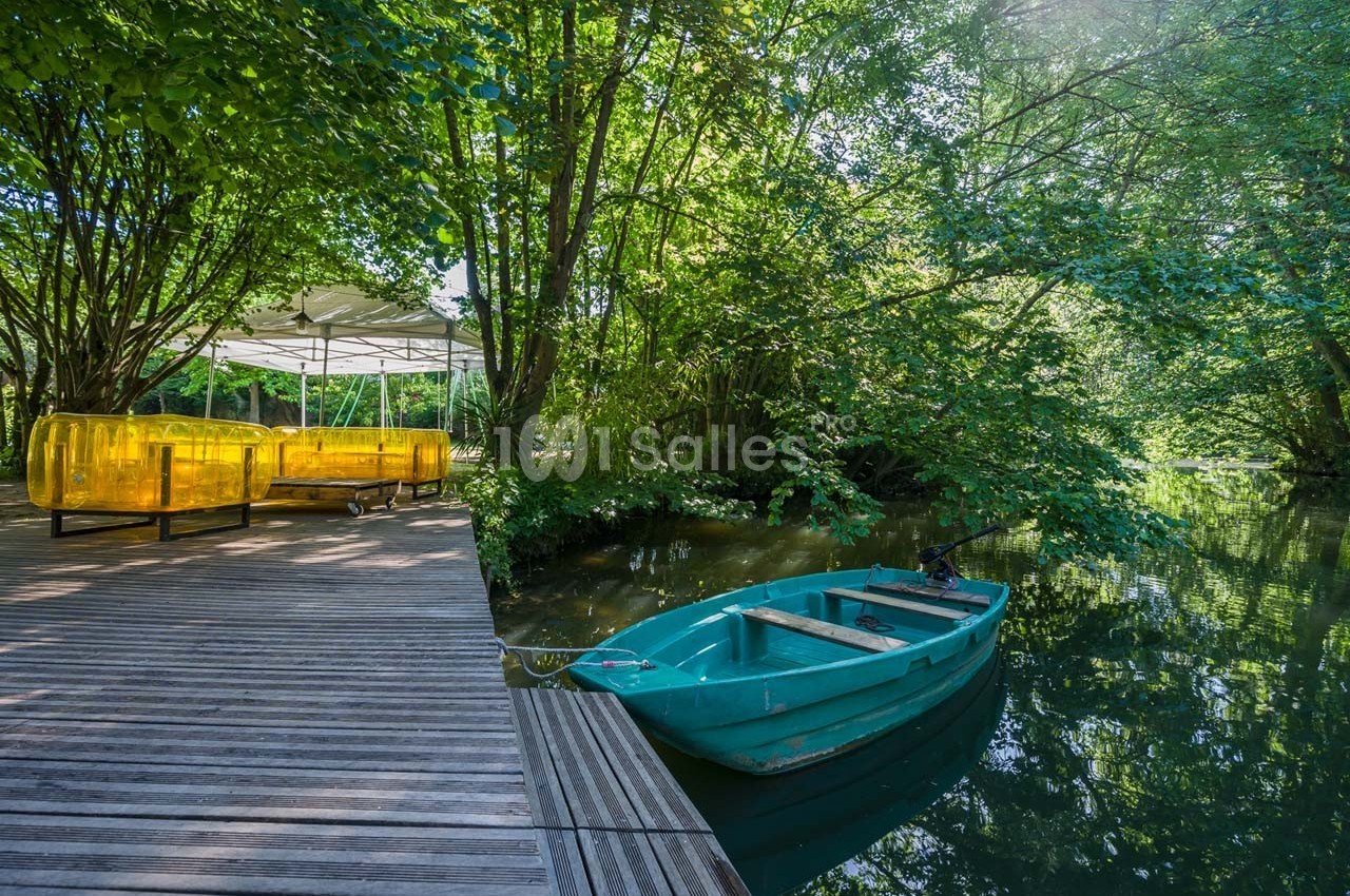 Barque bleue amarrée près d'un ponton en bois, entourée de végétation luxuriante et d'un coin détente avec des sièges jaunes.