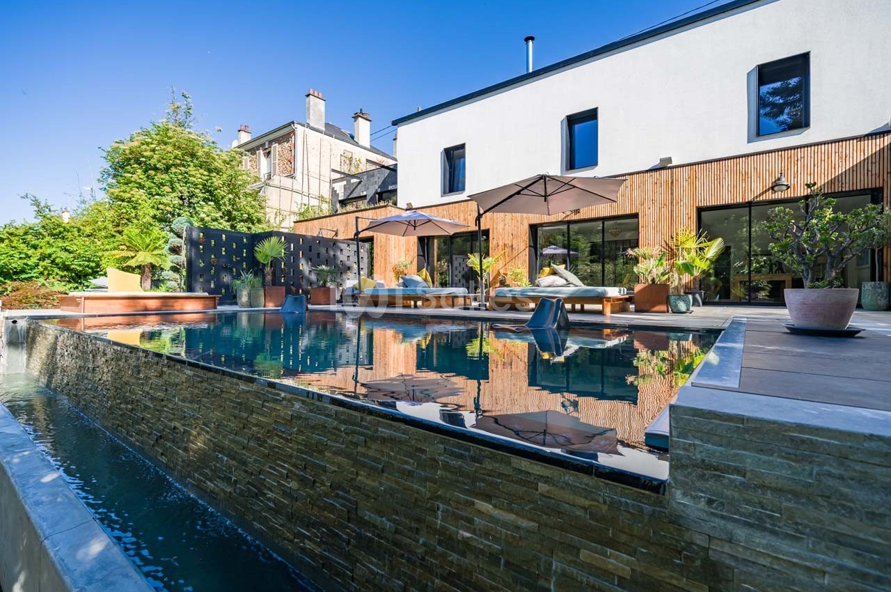 Piscine à débordement entourée de végétation et terrasses aménagées devant une maison moderne.
