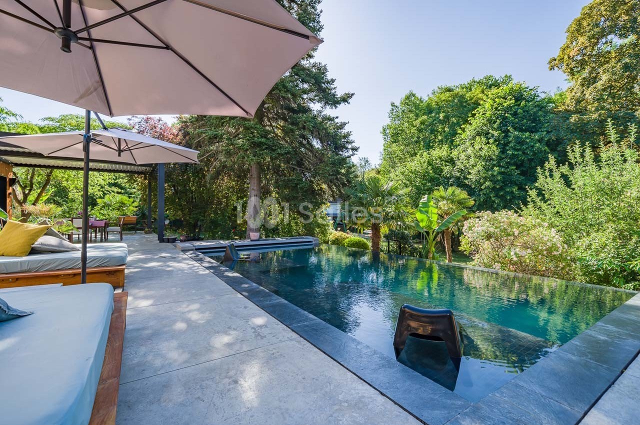 Piscine à débordement entourée de végétation luxuriante, avec transats et parasols sur une terrasse en pierre.