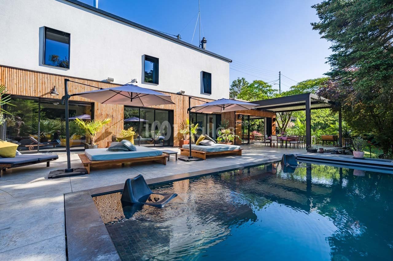 Terrasse moderne avec piscine, transats, parasols et végétation environnante devant une maison contemporaine.