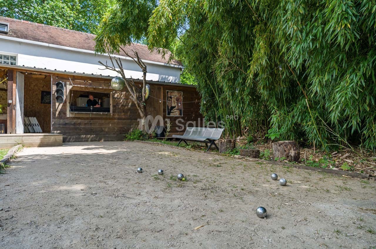 Terrain de pétanque en plein air avec des boules éparpillées, bordé de bambous et d'un bâtiment en bois.
