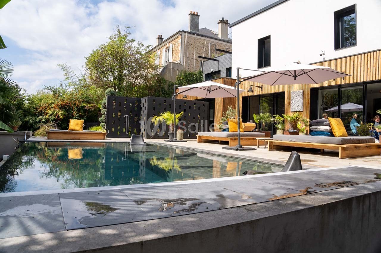 Piscine moderne entourée de terrasses en bois, mobilier extérieur et végétation, devant une maison contemporaine.