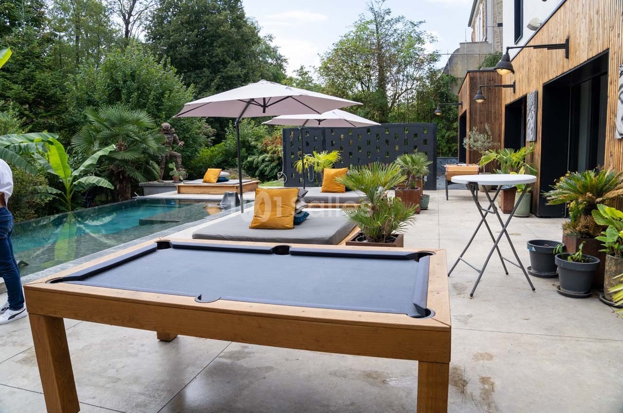 Terrasse avec piscine, table de billard, transats avec coussins jaunes, parasols et végétation environnante.
