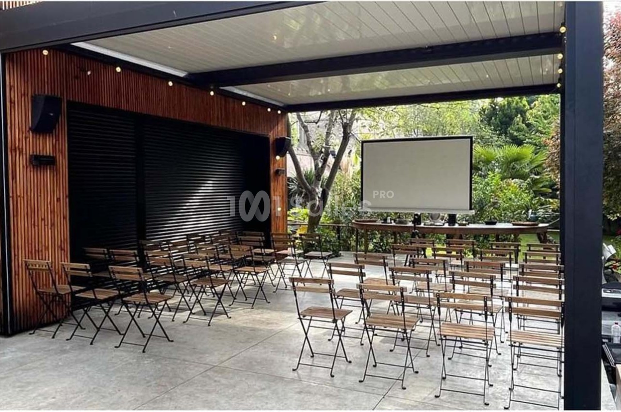 Salle extérieure aménagée avec des chaises pliantes, un écran de projection et un cadre verdoyant en arrière-plan.
