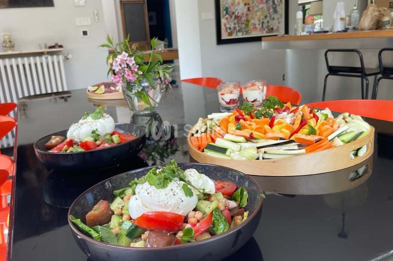 Assiette de salade colorée avec burrata au premier plan, plateau de crudités et fleurs en arrière-plan sur une table noire.