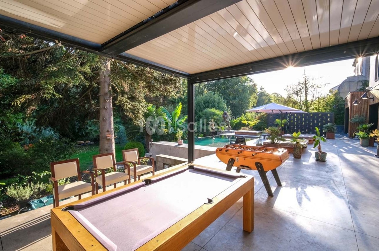 Terrasse aménagée avec billard, baby-foot, chaises et végétation environnante sous une pergola ouverte.
