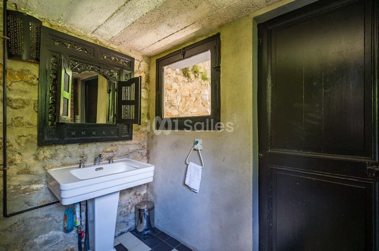 Salle de bain rustique avec lavabo, miroir encadré, mur en pierre, fenêtre et porte en bois sombre.