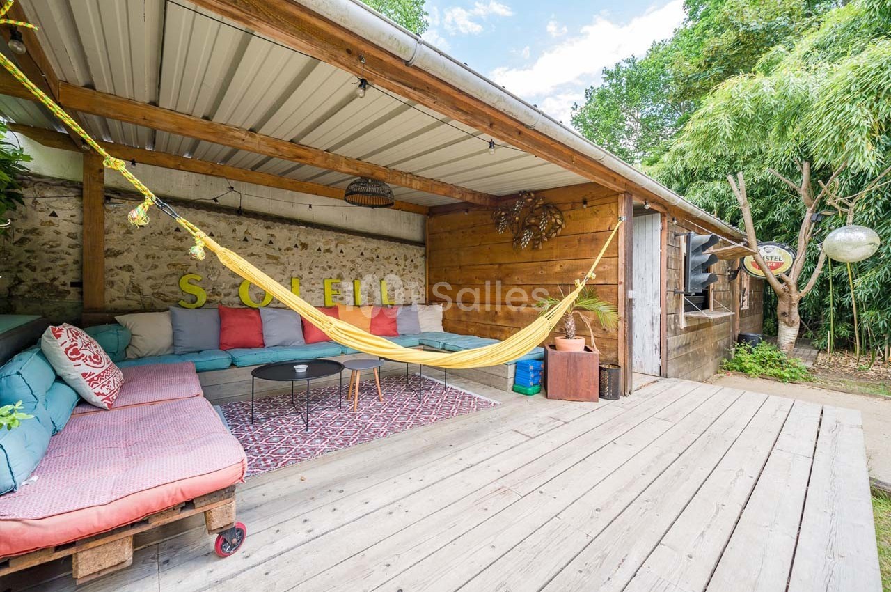 Espace extérieur couvert avec canapé en palettes, hamac jaune, tapis coloré et végétation environnante.