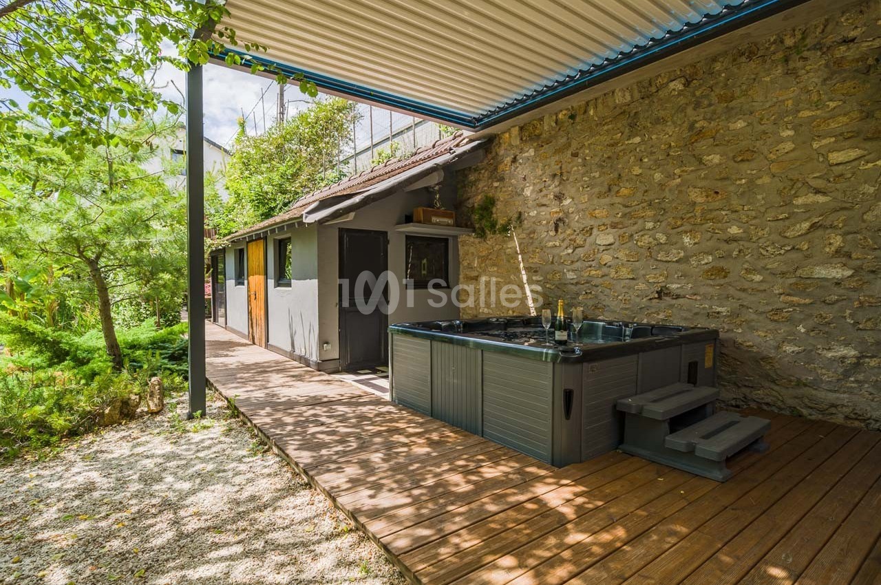 Terrasse en bois avec un jacuzzi, adossée à un mur en pierre, entourée de verdure et partiellement couverte.