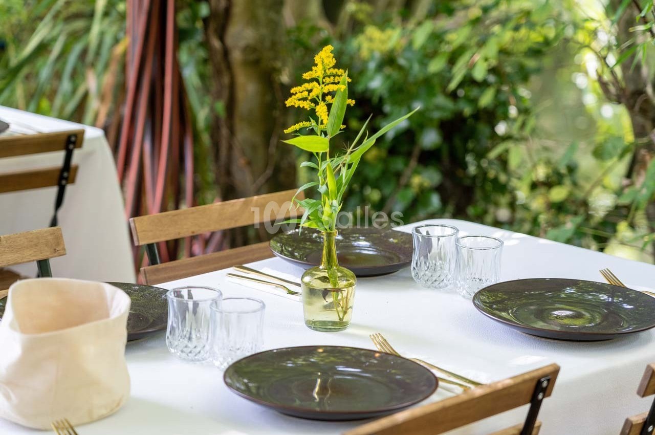 Table dressée avec assiettes noires, verres, couverts dorés et un vase contenant une fleur jaune, en extérieur verdoyant.