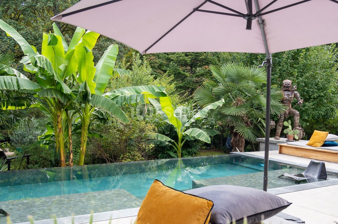 Piscine entourée de végétation tropicale avec un parasol et des coussins au premier plan.