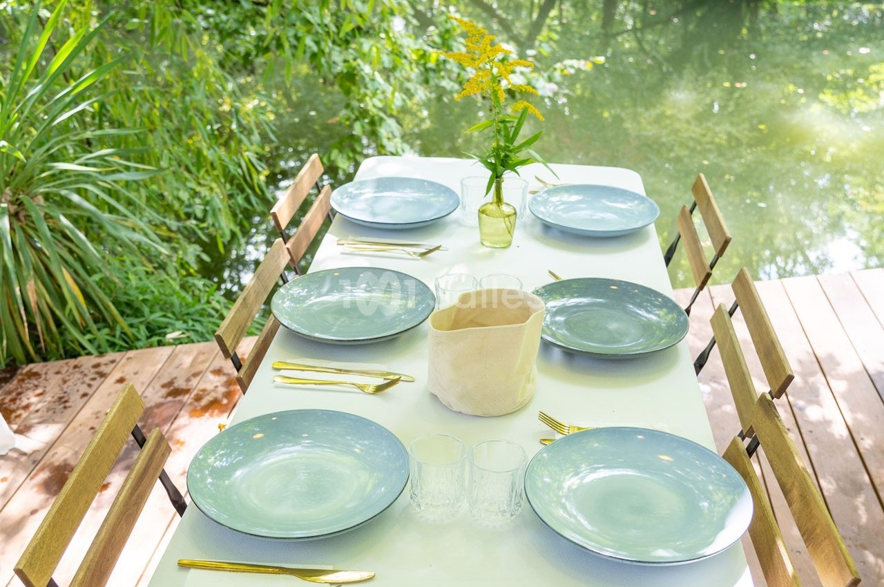 Table dressée en extérieur avec vaisselle bleue, couverts dorés, vase de fleurs jaunes et vue sur un cours d'eau.