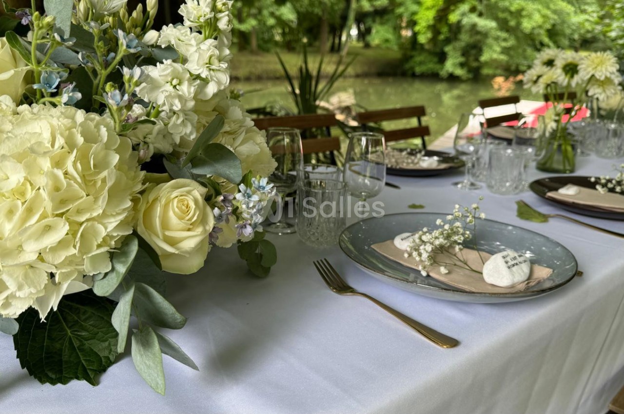 Table élégamment dressée en extérieur avec vaisselle raffinée, fleurs blanches et vue sur un étang verdoyant.