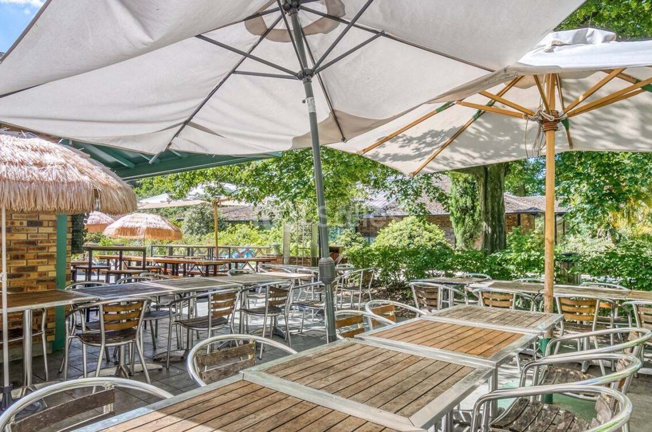 Terrasse extérieure avec tables et chaises en bois et métal, ombragée par des parasols et entourée de verdure.