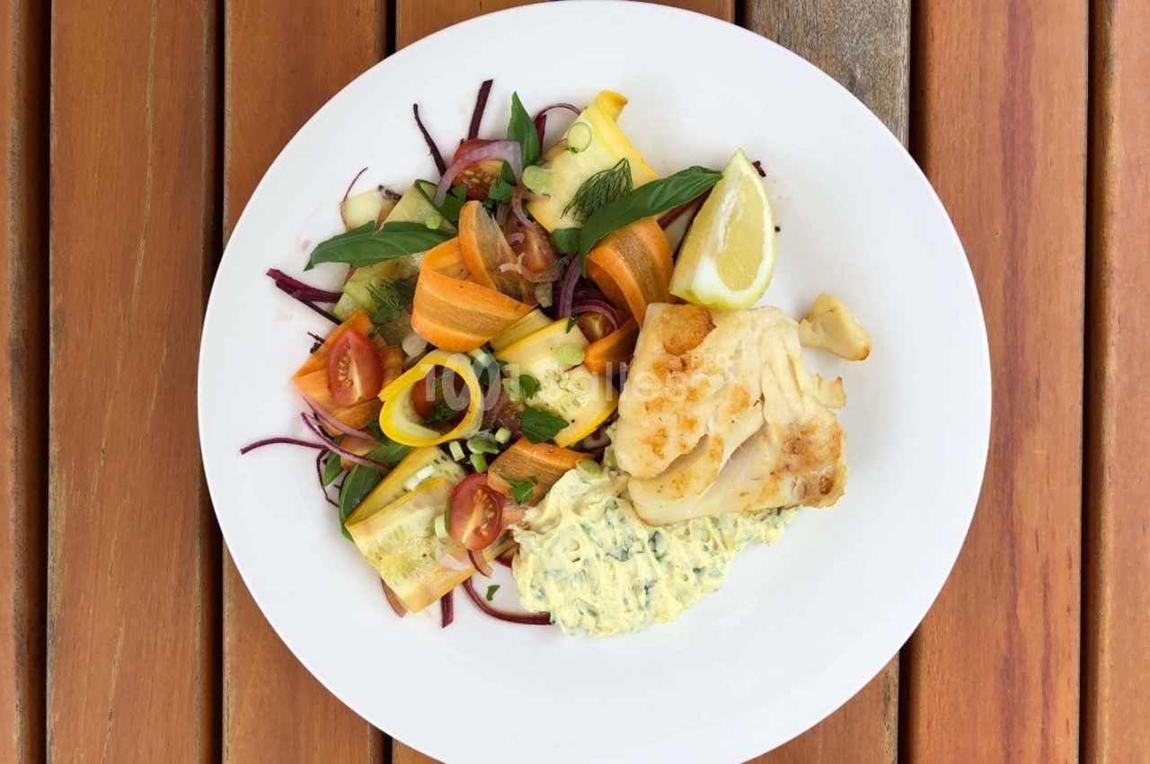 Assiette contenant du poisson grillé, une salade de légumes colorés et une sauce crémeuse, servie sur une table en bois.