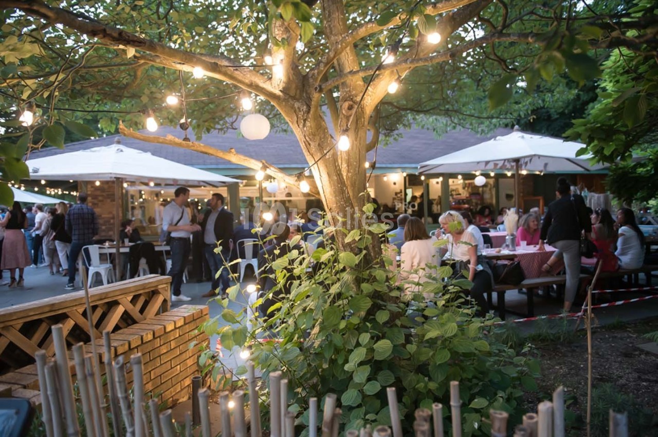 Terrasse animée en soirée avec des guirlandes lumineuses, des tables occupées et un arbre au premier plan.