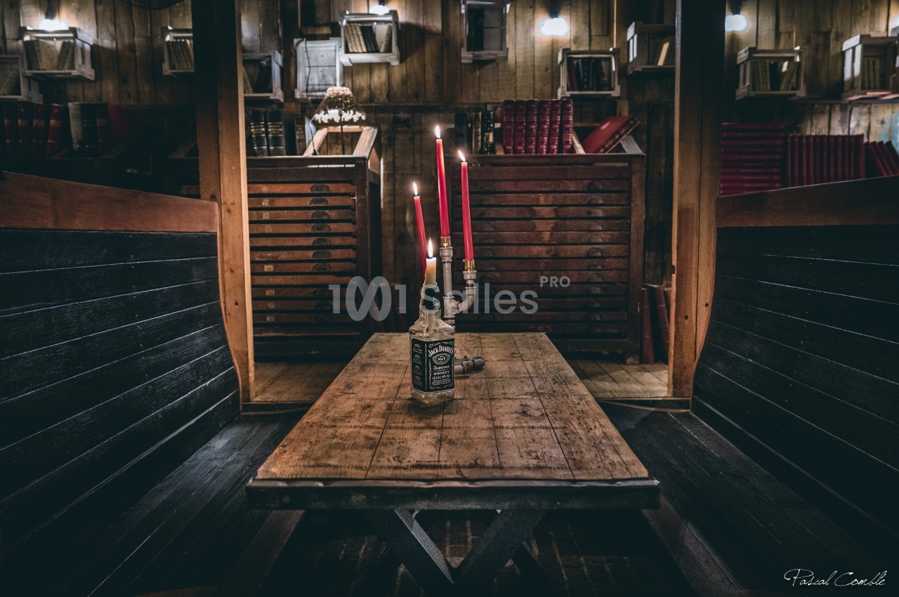Table en bois avec une bouteille transformée en chandelier à trois bougies rouges, dans un intérieur rustique avec étagères.